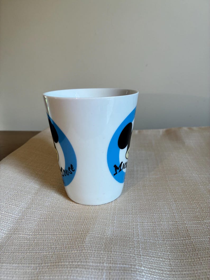 May include: A white ceramic mug featuring two blue circular graphics, each containing a black silhouette of Mickey Mouse's head and the text "Mickey Mouse" in black script. The mug has a smooth, tapered cylindrical shape with a wide rim.