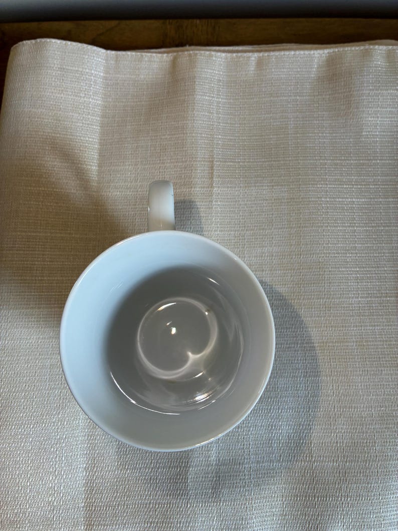 May include: A plain white ceramic mug viewed from directly above, showing a smooth, circular interior basin and a single curved handle extending from the side. The mug has a clean, minimalist design with a glossy finish and no patterns or text.