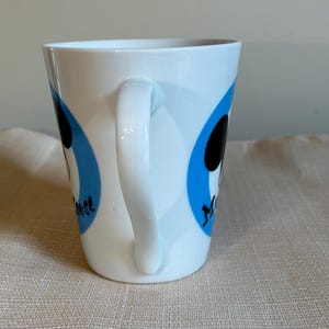 May include: A white ceramic mug featuring a curved handle and two blue circular graphics. Each blue circle contains a black silhouette of Mickey Mouse and the black text Mouse printed in a stylized font on the side of the mug.