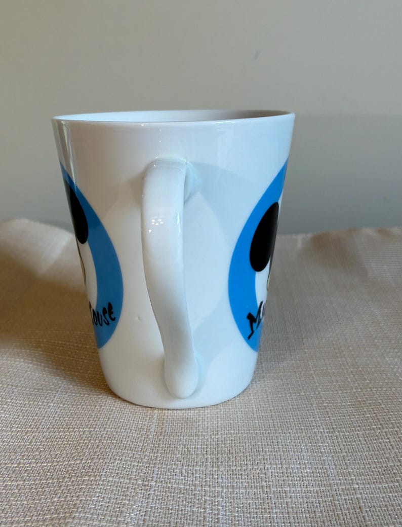 May include: A white ceramic mug featuring a curved handle and two blue circular graphics. Each blue circle contains a black silhouette of Mickey Mouse and the black text Mouse printed in a stylized font on the side of the mug.