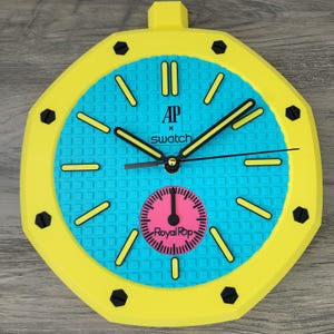 May include: A bright yellow octagonal wall clock with a textured turquoise dial. The face features the text AP x swatch and a pink circular sub-dial with the text Royal Pop. It has black hands and yellow hour markers with black screw accents.