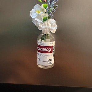 Könnte beinhalten: Ein durchsichtiges Glasfläschchen für Humalog Insulin-Lispro-Injektionen, das als dekorativer Magnet umfunktioniert wurde. Das Fläschchen hat ein weißes Etikett mit rotem Text und enthält ein Arrangement aus künstlichen weißen Blumen, einer kleinen rosa Blume und blauen Zweigen.