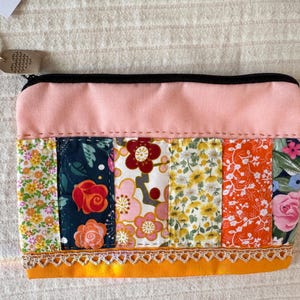 May include: A rectangular zipped pouch featuring a solid light pink upper section and a lower section composed of vertical fabric strips with various floral patterns in yellow, orange, red, and dark blue, finished with a gold lace trim.