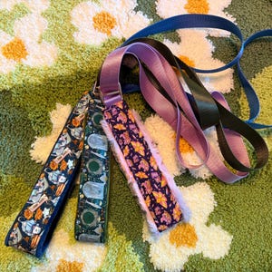 May include: A collection of three pet leashes featuring distinct patterns: one with blue and orange rat illustrations, one with green tombstone motifs, and one with a purple floral print. Each leash includes a soft, light purple faux fur lining.