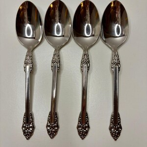 May include: A set of four identical silver-toned metal spoons arranged vertically. Each spoon features a polished oval bowl and a handle decorated with ornate, raised scrollwork patterns at the neck and base, separated by a smooth, flat center.