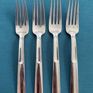 May include: A set of four identical silver-toned metal forks arranged in a vertical row. Each fork features a long, slender handle with a subtle, recessed vertical groove running down the center and four tines at the top end of the utensil.