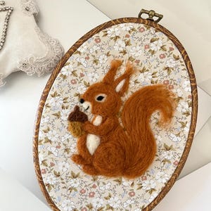 Op de afbeelding: Een ovale houten borduurring met een naaldgevilte roodbruine eekhoorn die een bruine eikel vasthoudt. De eekhoorn is geplaatst tegen een lichte stoffen achtergrond met een herhalend patroon van kleine witte en roze bloemetjes.