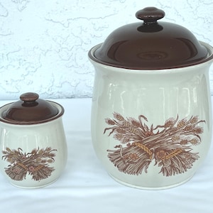 May include: Two ceramic canisters with glossy cream-coloured bodies and dark brown lids. Each canister features a brown graphic illustration of a tied wheat sheaf on the front. The larger canister is positioned to the right of the smaller one.