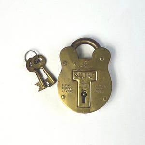 May include: A solid brass Squire padlock with a rounded body and a shackle, accompanied by two matching keys on a metal ring. The lock features engraved text including "Squire," "Four Brass Levers," and "Genuine Solid Brass" on its front surface.