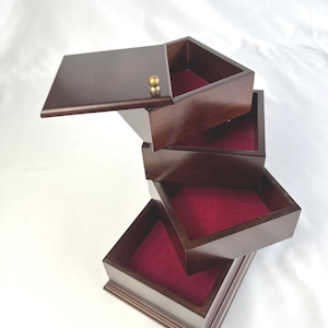 May include: A dark brown wooden jewellery box with a tiered, rotating design featuring four square compartments. Each compartment is lined with deep red velvet fabric. The top lid includes a small, round, metallic gold-coloured knob handle.