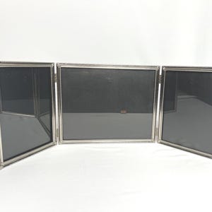 May include: A silver-toned metal triptych photo frame featuring three rectangular panels connected by hinges. Each frame has a thin, beaded border design surrounding a black insert. The structure is designed to stand upright on a flat surface.