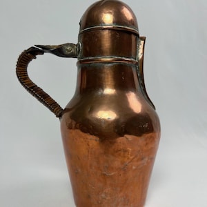 May include: A copper pitcher with a rounded body, a domed lid featuring a small knob, and a curved handle wrapped in dark brown textured material. The metal surface shows a polished finish with subtle variations in color and texture throughout.