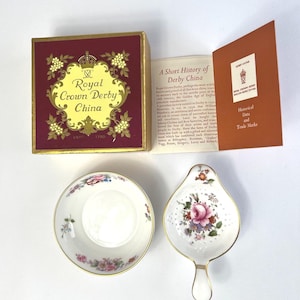May include: A Royal Crown Derby bone china set comprising a round bowl and a handled strainer, both decorated with floral motifs and gold rims. The set is accompanied by a burgundy and gold presentation box and a booklet titled A Short History of Derby China.