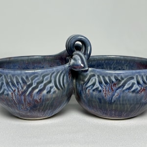 May include: A double-bowl ceramic server featuring a glossy glaze in shades of blue, purple, and hints of red. The two bowls are connected by a central handle with a decorative curled loop at the top, and the exterior surfaces feature a textured, wavy pattern.