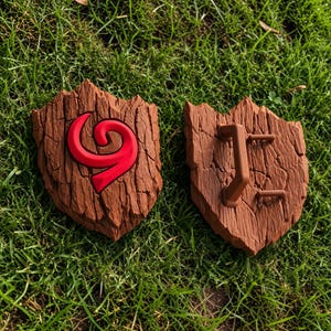 Pode incluir: Dois objetos em forma de escudo castanhos com uma superfície texturada semelhante a casca de árvore. O escudo esquerdo apresenta um emblema em espiral vermelho em relevo, enquanto o direito mostra duas pegas castanhas fixadas na parte traseira.