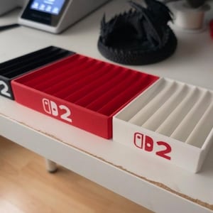 Puede incluir: Tres bandejas de almacenamiento rectangulares en negro, rojo y blanco, cada una con múltiples ranuras paralelas para organización. El frente de cada bandeja muestra un logotipo impreso con un icono de mando seguido del número 2.