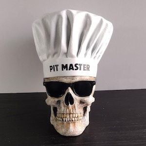 Peut inclure: Une réplique de crâne humain portant des lunettes de soleil noires et une toque de chef blanche. La toque présente une large bande avec le texte PIT MASTER imprimé en lettres capitales noires. Le crâne a une finition blanc cassé vieillie.
