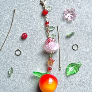 May include: A beaded charm featuring a gold-tone lobster clasp, a red bead, small green leaf beads, a pink flower-shaped bead, and a round orange and red fruit-shaped bead. Loose components include two metal pins, two rings, and beads.