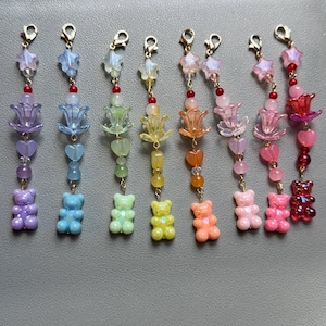 May include: Eight colorful beaded charms featuring a gold-tone lobster clasp, a star-shaped bead, a flower-shaped bead, a heart-shaped bead, and a gummy bear-shaped pendant at the bottom. The charms are arranged in a row in shades of purple, blue, green, yellow, orange, light pink, and red.