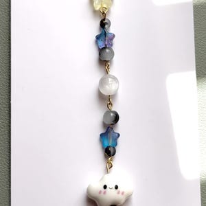 May include: A beaded charm featuring a gold-tone metal clasp, a translucent yellow star, a blue star, a white sphere, and a second blue star. The charm ends with a white ceramic cloud bead with a smiling face, pink cheeks, and small grey beads.