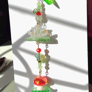 May include: A beaded charm featuring a gold-tone lobster clasp, a translucent green leaf-shaped bead, small light green and red round beads, a clear flower-shaped bead, a star-shaped bead, and a red mushroom-shaped bead with a green base.