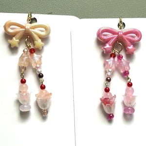 May include: Two dangle earrings featuring a bow at the top with two hanging strands. The left earring has a cream bow, star charms, and pink and red beads. The right earring has a pink bow, star charms, and pink and red beads with flower-shaped charms.