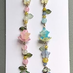 May include: Two long beaded charms featuring gold-tone lobster clasps, star-shaped beads, and various floral and fruit-shaped accents. The left charm includes pink and white beads with a pink strawberry, while the right features blue and yellow beads with blue flower-shaped charms and green leaf accents.