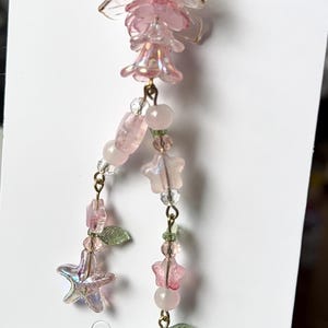 May include: A decorative charm featuring a clear glass bell-shaped top, pink flower-shaped beads, translucent star-shaped beads, and a small pink fruit-shaped bead with green leaves. The components are connected by gold-toned metal links.