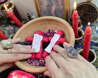 May include: A terracotta bowl containing two red fabric cross-shaped dolls, dried rose petals, and two white paper slips with handwritten text. The bowl is surrounded by four lit red candles, a gold-framed picture, and a circular metal talisman.