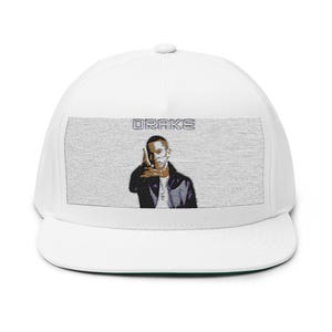 May include: A white snapback cap featuring a rectangular patch on the front panel. The patch displays a graphic illustration of a person pointing forward with the text DRAKE printed in a block font above the figure in a grey color scheme.