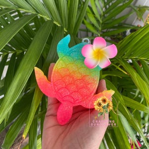 May include: A colorful, glittery turtle-shaped ornament with a gradient of blue, yellow, orange, and pink. A pink and yellow flower is attached to the turtle's head. The ornament is held in a hand against a backdrop of green leaves.