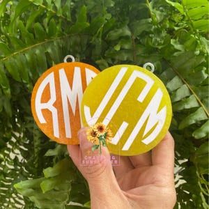 May include: Two round, personalized keychains. One is orange with white initials "RM", the other is yellow with white initials "JCM". Both have a silver loop at the top. The keychains are held in front of a background of green leaves.