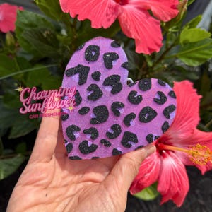 May include: A heart-shaped car freshener featuring a vibrant purple base with a black glittery leopard print pattern. The surface has a textured, sparkling appearance, and the item is shaped like a classic heart with a rounded top and pointed base.