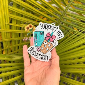May include: A glittery white acrylic ornament featuring a teal credit card, an iced coffee cup with a pink bow, and the black text Support my obsession. The design includes a small sunflower graphic and a pink flower accent on the surface.