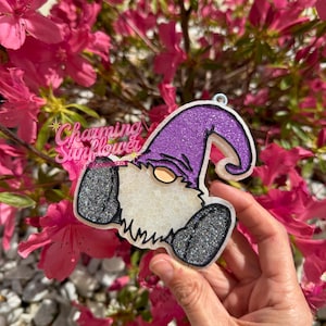 May include: A gnome-shaped ornament with a purple glitter hat, black glitter shoes, and a white beard. The ornament has a small metal loop for hanging. The text "Charming Sunflower" is visible in the upper left corner.