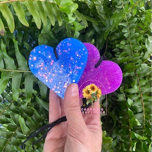 May include: Two heart-shaped air fresheners, one blue and one purple, with glitter accents. The blue heart has pink glitter, and the purple heart has silver glitter. Both have black string for hanging. A small sunflower graphic is visible.