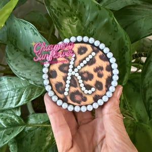 May include: A circular car freshener featuring a brown and black leopard print pattern. The letter K is formed by small white beads in the center, and the outer edge of the circle is bordered by a continuous row of white beads.