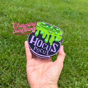 May include: A black cauldron-shaped car air freshener featuring the text HOCUS POCUS in purple lettering. The top of the cauldron has a bright green, textured, dripping liquid design accented with small, round, light green beads throughout.