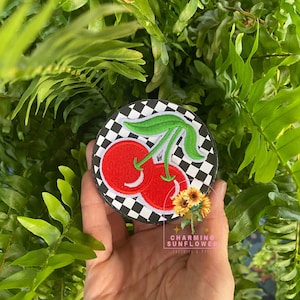 May include: A round patch featuring a pair of red cherries with green stems and leaves, set against a black and white checkered background. The patch has a black border and the text "CHARMING SUNFLOWER" is visible.