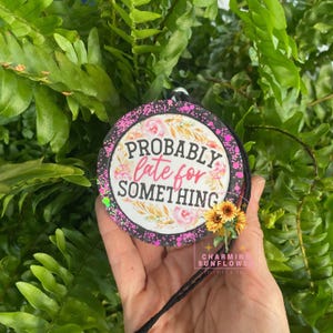 May include: A round hanging ornament with a black border featuring pink paint splatters. The center displays the text 'PROBABLY late for SOMETHING' surrounded by floral illustrations and a small attached artificial sunflower cluster.