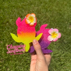 May include: Two sea turtle-shaped air fresheners with glittery finishes. One is a pink-to-yellow gradient, and the other is a purple-to-pink gradient. Each turtle features a textured shell pattern and a single white and yellow plumeria flower.