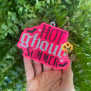 May include: A bright neon pink, irregularly shaped hanging ornament featuring the text HOT GHOUL SUMMER in black and silver glitter lettering. The design includes small black bat silhouettes, silver stars, and a small sunflower graphic.
