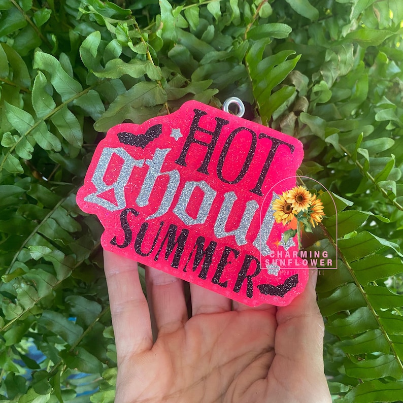 May include: A bright neon pink, irregularly shaped hanging ornament featuring the text HOT GHOUL SUMMER in black and silver glitter lettering. The design includes small black bat silhouettes, silver stars, and a small sunflower graphic.