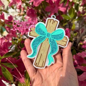 May include: A cross-shaped ornament featuring a gold glitter finish and a prominent teal glitter bow. The ornament has a white border and a small metal loop at the top for hanging. The design is flat with a textured, sparkling appearance.