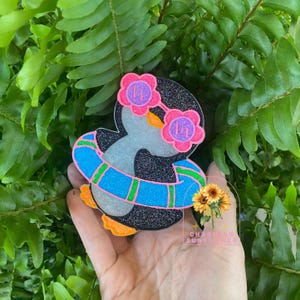 May include: A whimsical penguin-shaped accessory with a glittery black body, wearing pink flower-shaped sunglasses and a blue and green striped floatie. The penguin has orange feet and is set against a backdrop of green foliage.