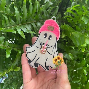May include: A ghost-shaped ornament featuring a glittery pink beanie with a brown patch, holding a brown iced coffee cup with a pink straw. The ghost has black eyes and a white body with black outlines, and a small metal loop at the top for hanging.