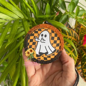 May include: Round Halloween ornament with a white ghost graphic giving the middle finger, set against an orange and black checkered background. The ornament has a glittery orange and black border, with a small sunflower bouquet and the text "Charming Sunflower".