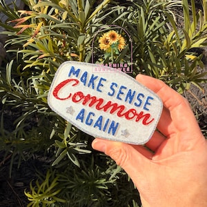 May include: A rectangular glittery white plaque with rounded corners featuring the blue text MAKE SENSE and AGAIN, with the red glittery text Common in the center. Two small silver star shapes are positioned below the word Common.