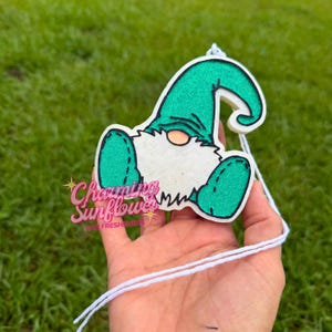 May include: A hanging air freshener shaped like a gnome with a glittery green hat and boots, a white beard, and a peach-colored nose. The design features a black outline and includes a white string for hanging. The text Charming Sunflower Car Fresheners is printed in pink and white script over the lower portion of the gnome figure.
