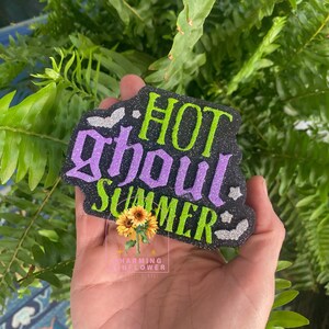 May include: A black glittery sign with the text HOT GHOUL SUMMER in lime green and purple glittery letters. The sign features small silver glittery bat and star accents and a central graphic of two yellow sunflowers with green leaves.